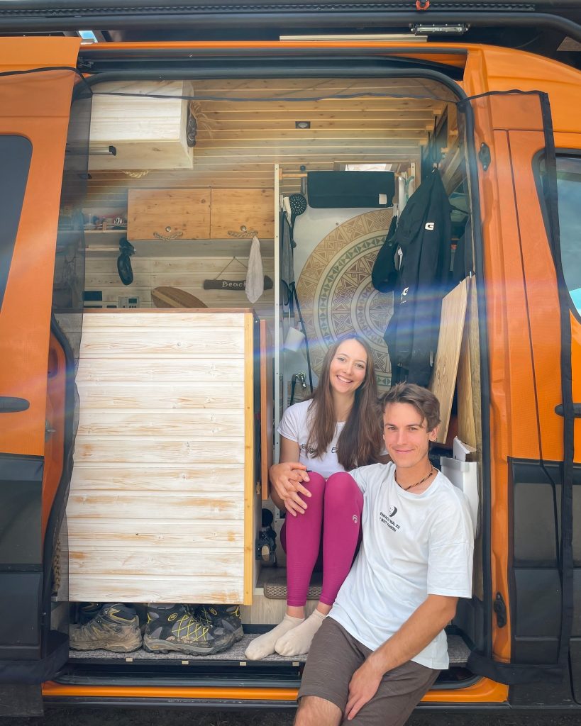 Patricia und Joel sitzen in der offenen Schiebetür ihres selbstausgebauten orangenen Sprinter Campervan