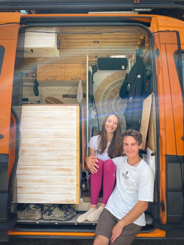 Patricia und Joel sitzen in der offenen Schiebetür ihres selbstausgebauten orangenen Sprinter Campervan